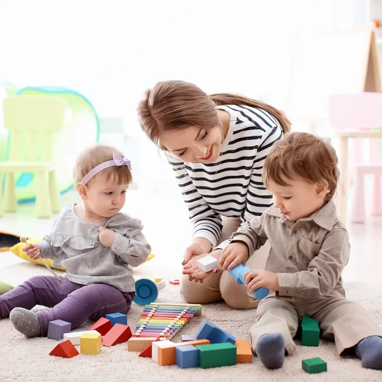 Junge Frau spielt mit zwei kleinen Kindern am Fussboden - Babysitter:in